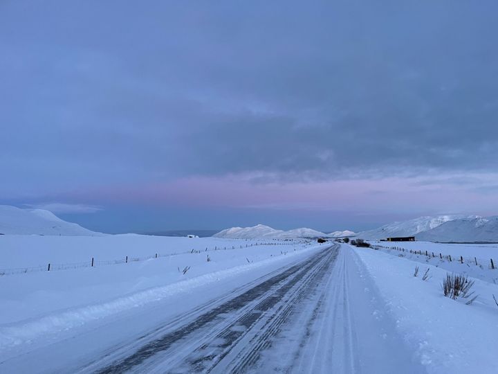 The road close to my dad's place. Looking north. The road is covered in snow and ice. It is twilight and the sky is mostly blue but kind of purple at the top. Snow capped mountains are visible across the fjord.